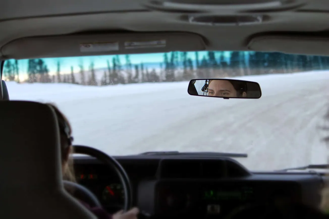 Frosty winter road viewed from inside a vehicle with a driver and a woman looking through the rearview mirror, snowy landscap