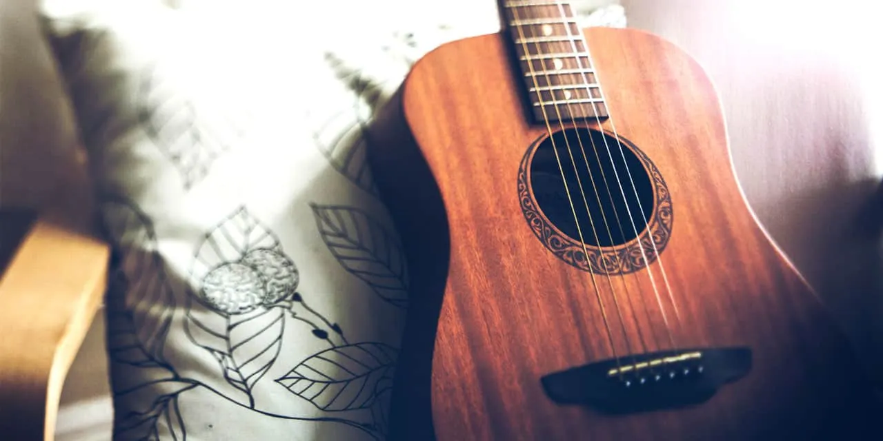 Guitar resting on bed with floral pillow, acoustic wood guitar with intricate rosette design, cozy musical instrument setting