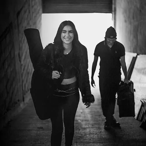 Young woman smiling with backpack and guitar case walking through an urban tunnel, representing music industry professionals