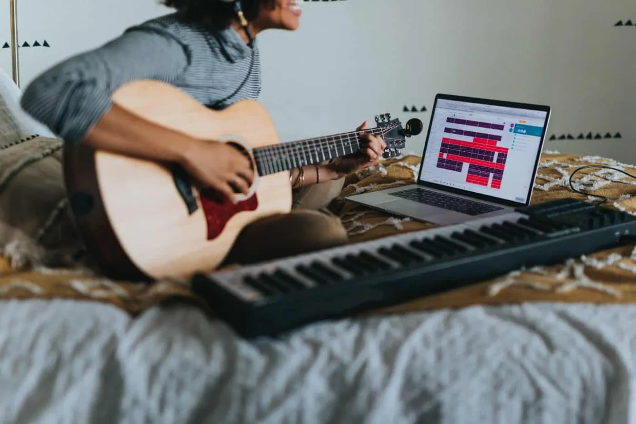 Guitarist creating music using a laptop, keyboard, and acoustic guitar in a home studio setup for digital music production an