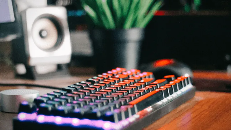 Keyboard with RGB lighting on a wooden desk, gaming setup with speakers and monitor in the background, ideal for digital musi