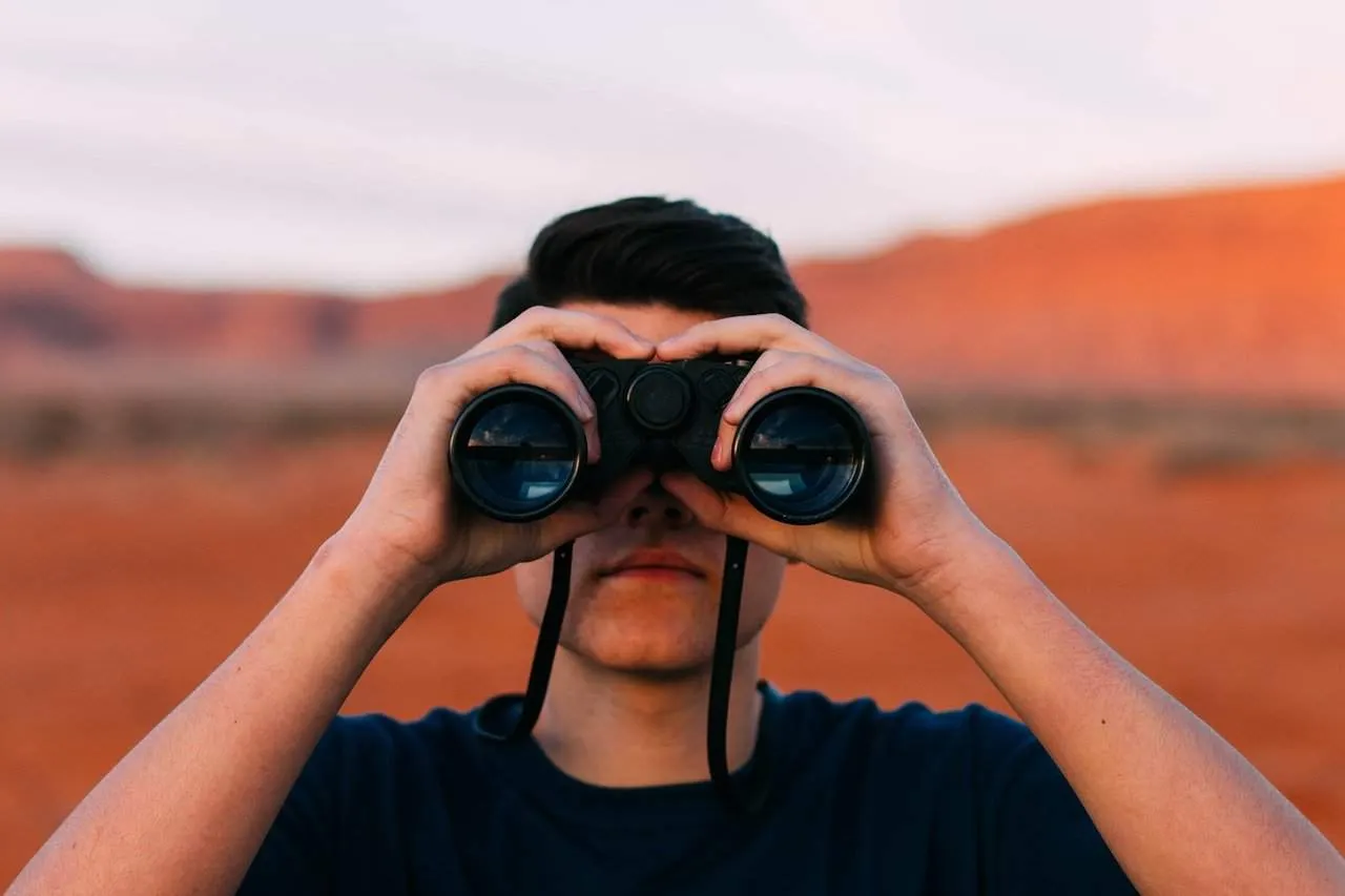 A young man looking through binoculars in a desert landscape with red rock formations in the background.