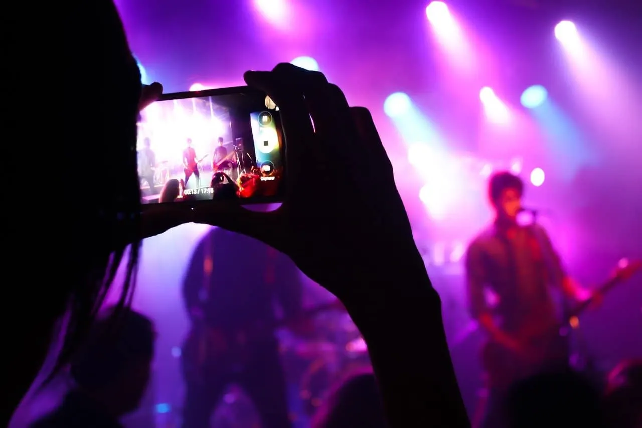 Wide-angle shot of a person recording a live music concert on smartphone with a band performing on stage illuminated by vibra