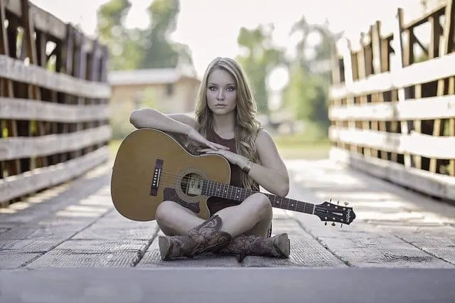 Acoustic guitar player woman sitting outdoors on wooden bridge with guitar, casual music artist, young female musician, singe
