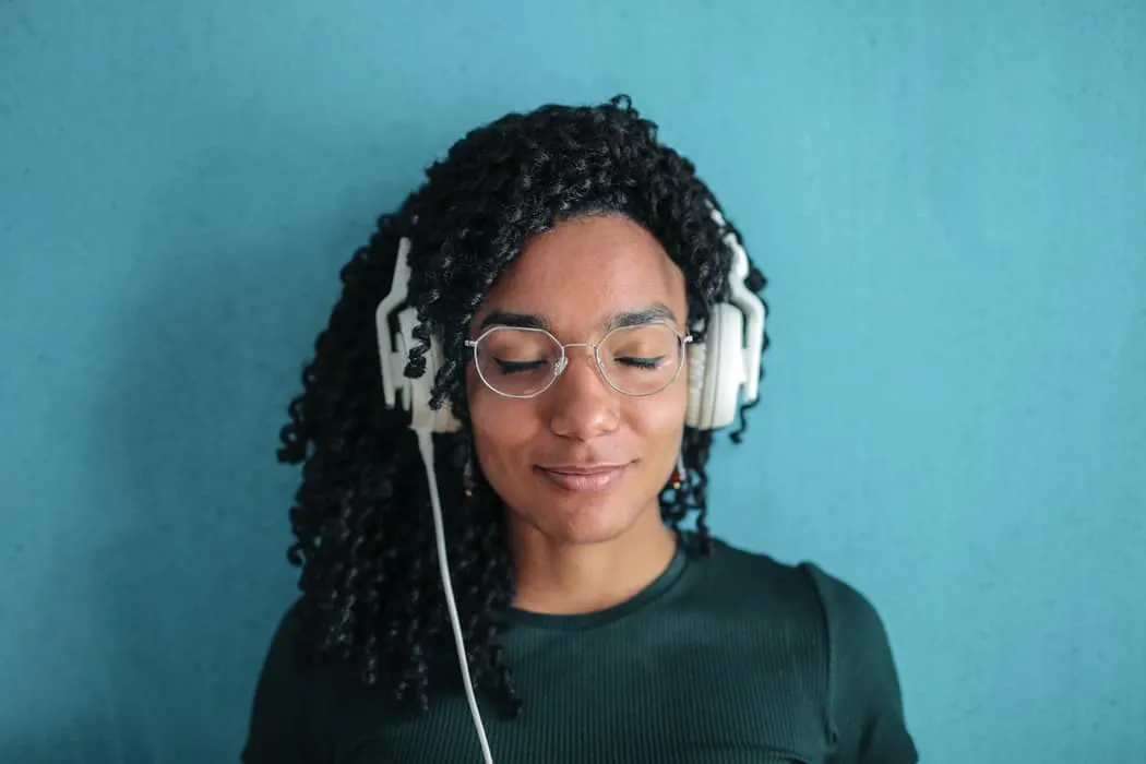 Relaxed woman listening to music with headphones, wearing glasses and a dark shirt, standing against a blue background.