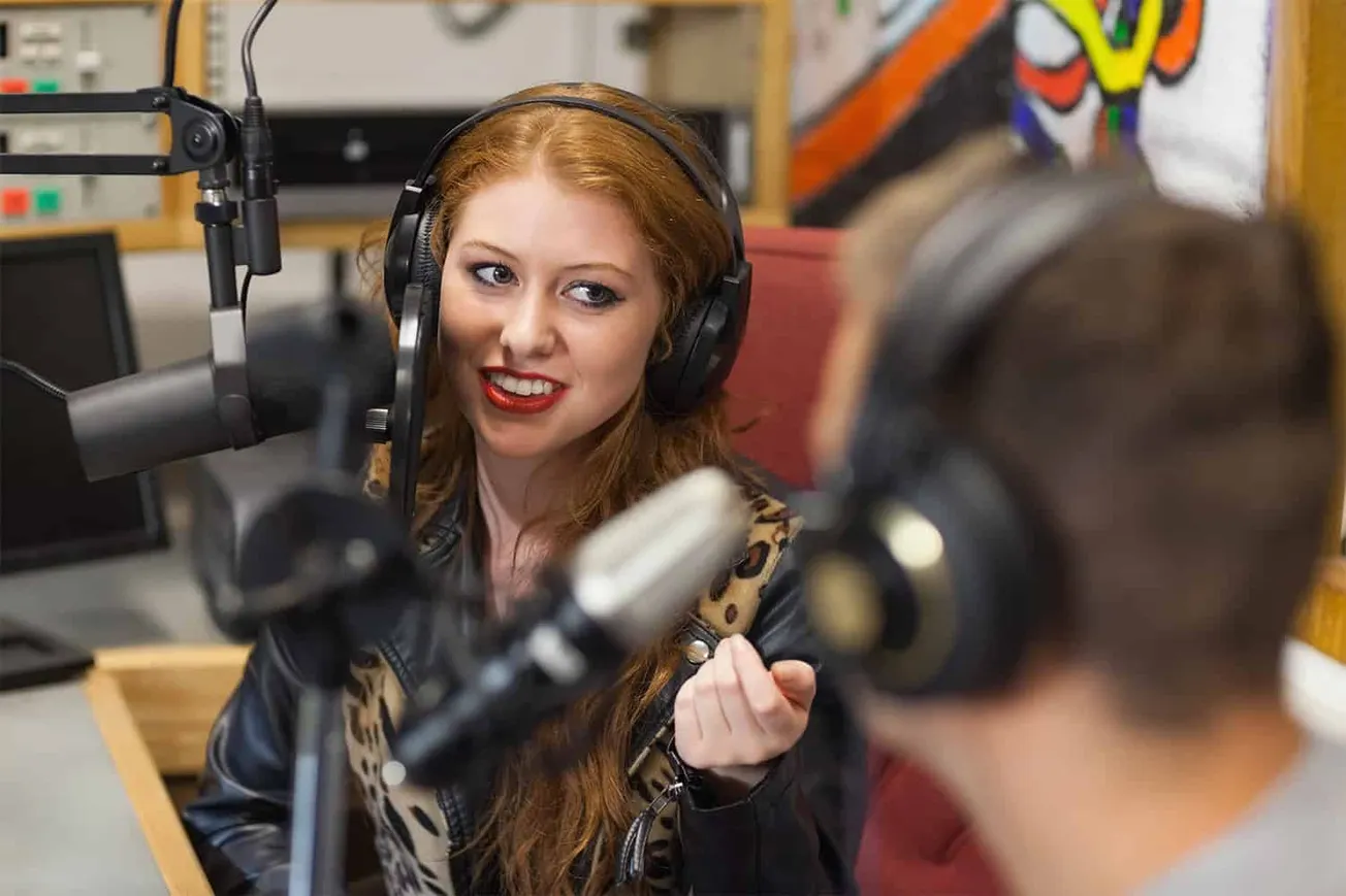 Female radio host speaking into microphone during podcast recording, wearing headphones, in a studio setting with colorful ar