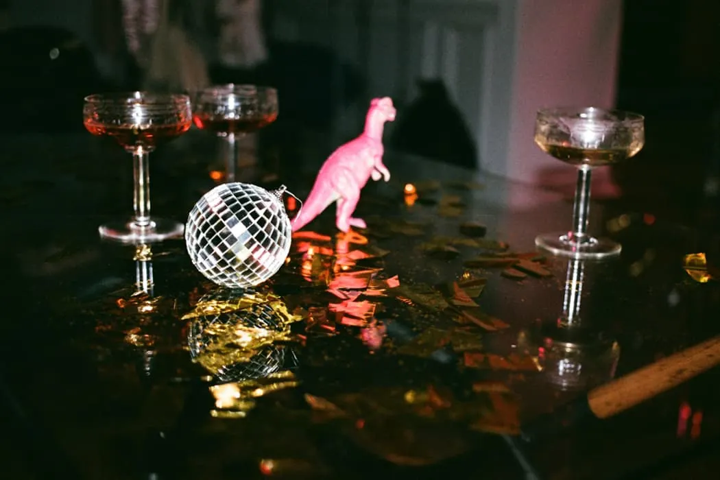 Colorful party scene with disco ball, pink dinosaur toy, and champagne glasses on a reflective surface, celebrating a fun nig