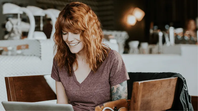 Young woman working on laptop at a cafe, smiling, with music equipment and cozy atmosphere in the background.