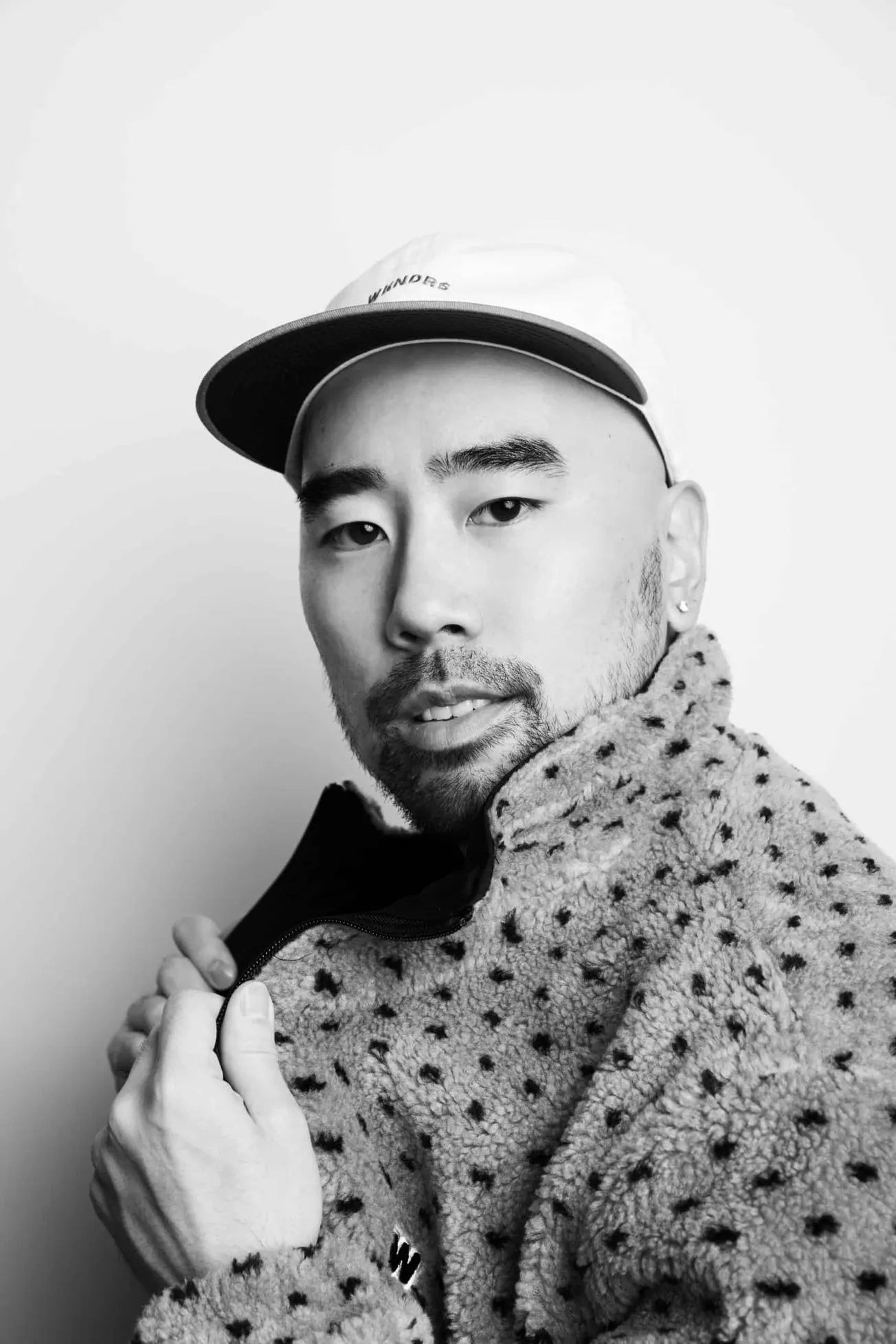 Youthful Asian man in a baseball cap and patterned fleece jacket, looking confidently at the camera against a plain backgroun