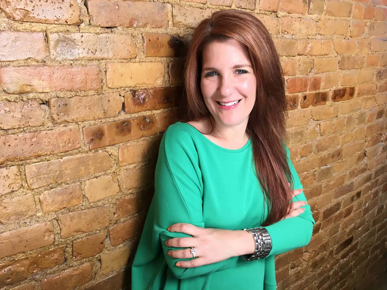 Friendly redhead woman smiling confidently against a rustic brick wall, wearing a bright green top and silver watch, represen