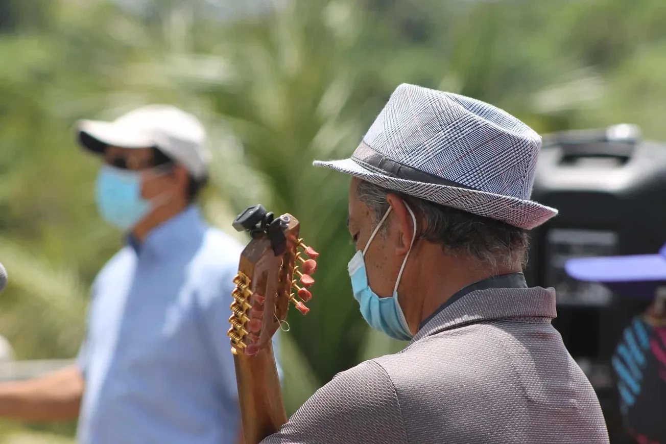 Vintage-style man playing guitar outdoors during daytime, wearing a checkered hat and face mask, with blurry person in backgr