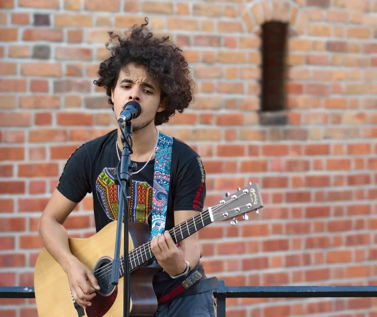 Young male musician performing live with guitar against brick wall background, singing into microphone, showcasing street mus