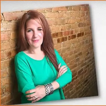 Young woman with red hair wearing a bright teal top, smiling confidently against a rustic brick wall background.