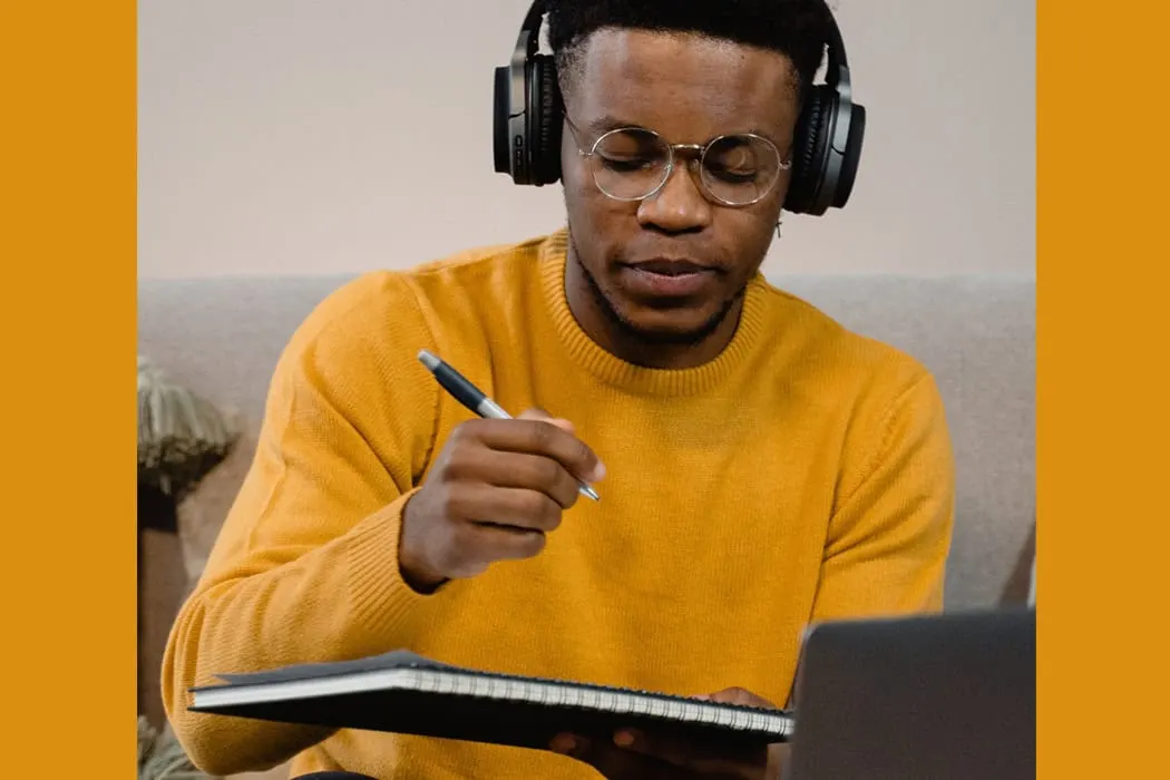 Young man with headphones writing in a notebook while working on a laptop, digital music creation, music production, remote w
