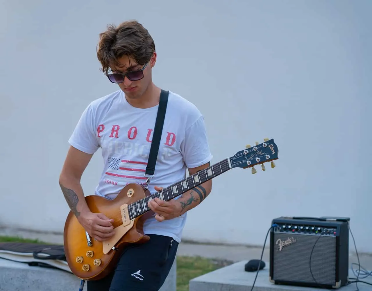 Guitarist playing outdoor music performance with amplifier, young man with sunglasses, casual white t-shirt, tattoos, electri