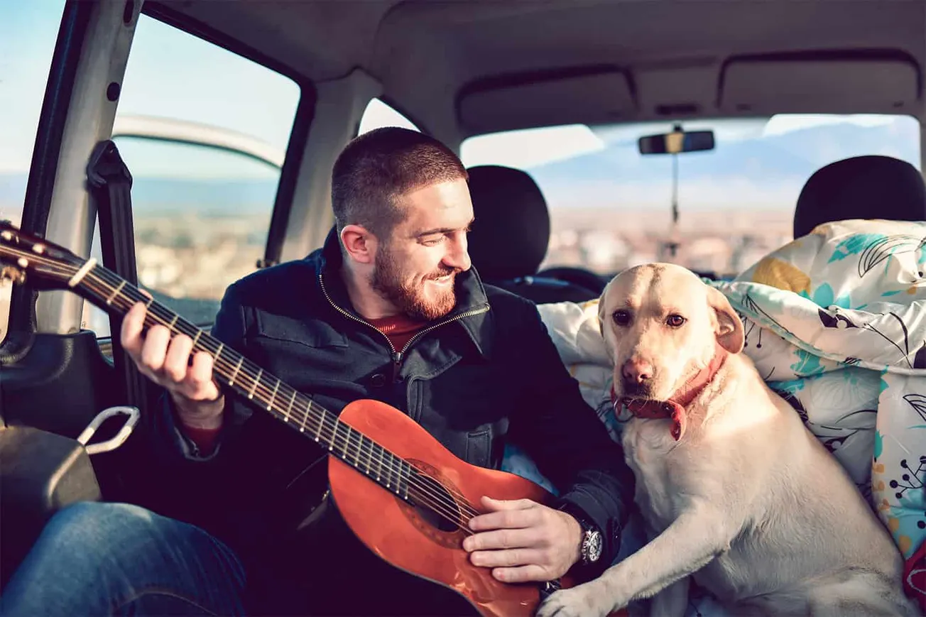 Guitar-playing man with a Labrador in the back of a van, enjoying music and outdoor adventure, promoting music content for Hy