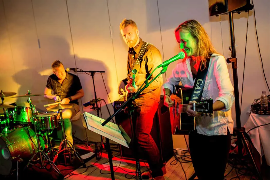 Hypebot, live musical performance featuring a guitarist and female singer with a band drummer in the background, vibrant stag