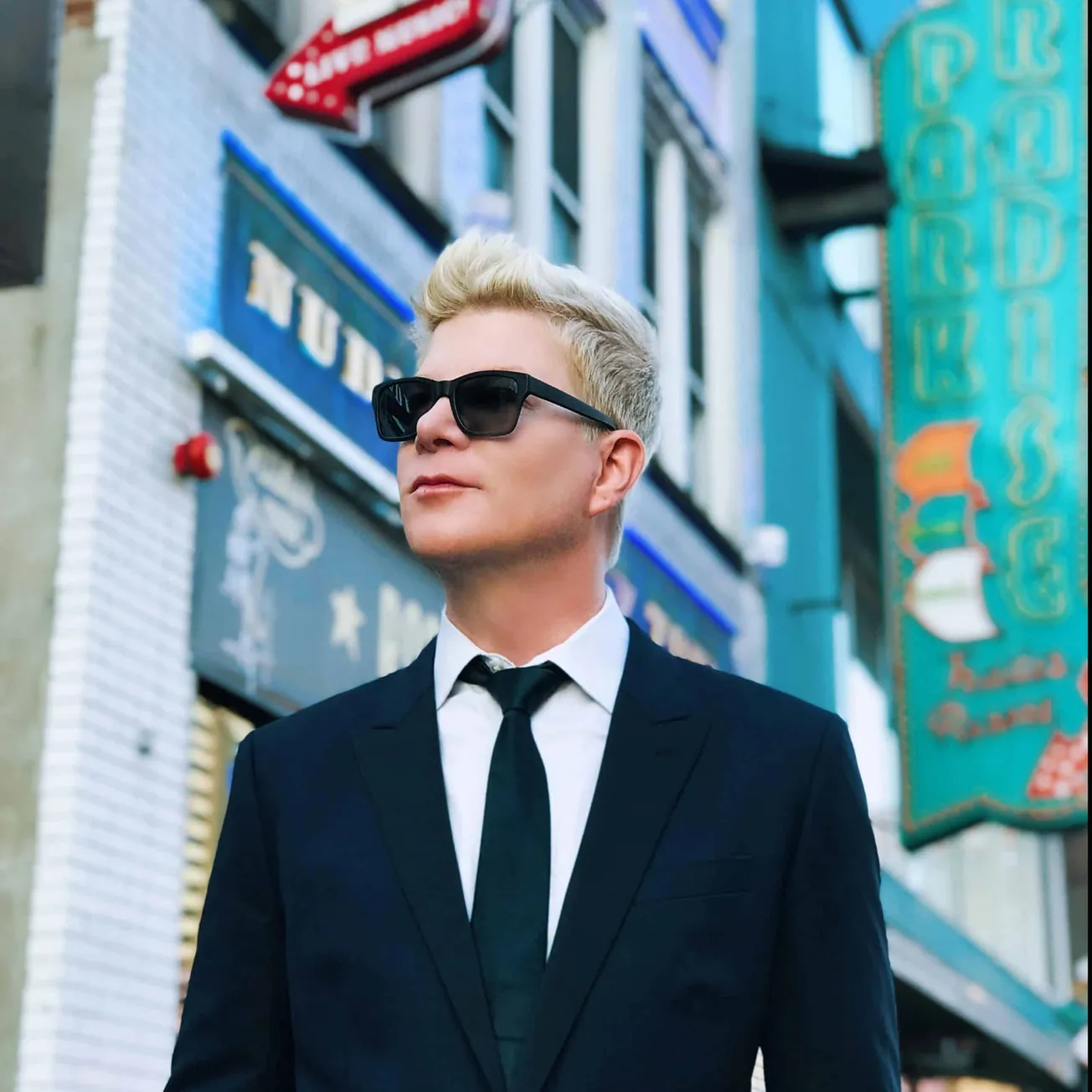 Stylish young man in a black suit and tie with sunglasses standing on an urban street with colorful neon signs, representing
