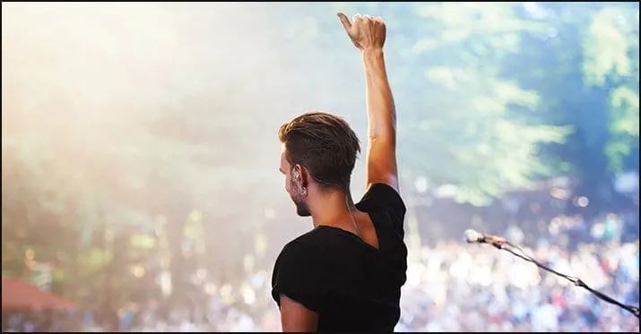 A concert performer raising a fist on stage with a large audience and lush green trees in the background.