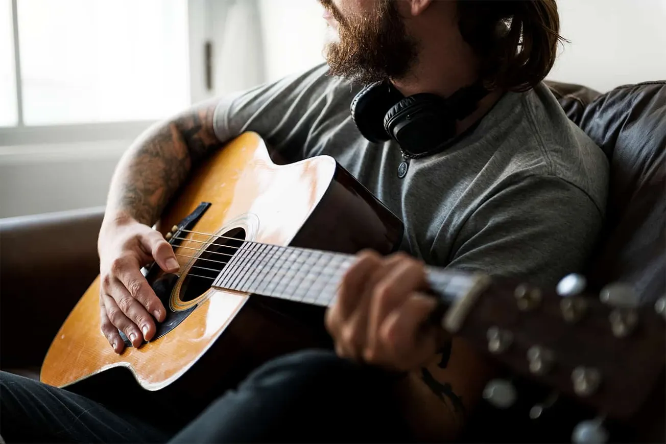 Guitar player with long hair and tattoos playing acoustic guitar on a couch, wearing headphones, in a bright indoor setting.