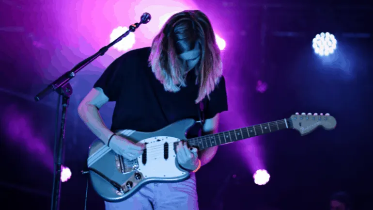 Electric guitar player performing live on stage under vibrant purple and blue lighting, showcasing indie or alternative music