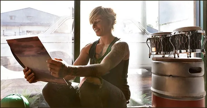 Young woman with tattoos reading a newspaper in a creative studio environment, with musical instruments around her, showcasin