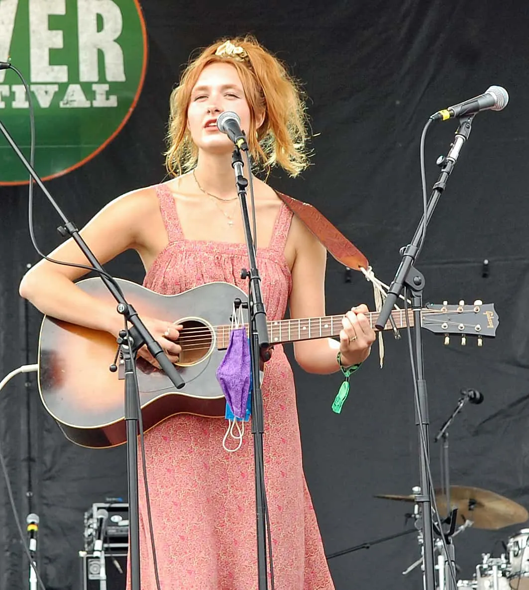 Female musician performing live with guitar at outdoor music festival, singing into microphone during a concert.