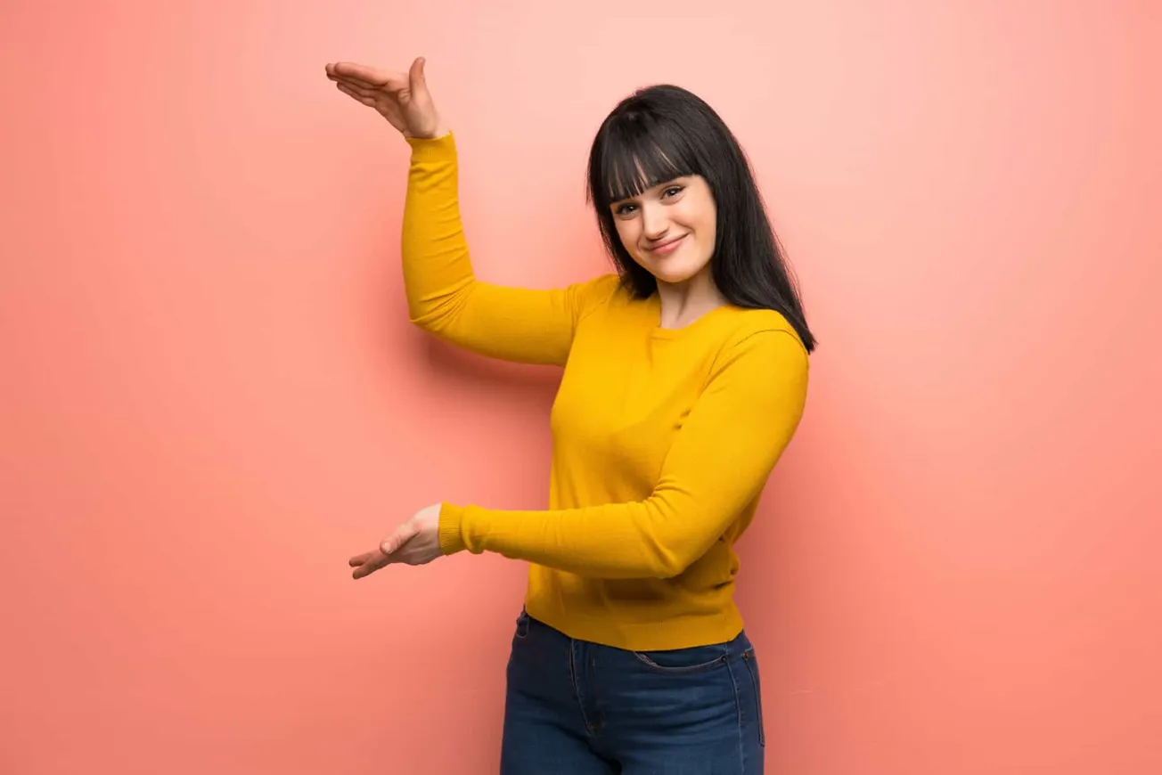 Bright young woman with black hair in a yellow sweater smiling and gesturing with her hands on a pink background.
