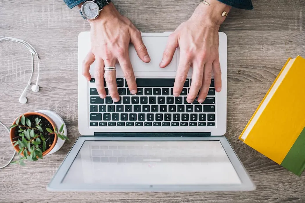 Hands typing on a silver laptop keyboard with books, headphones, and a potted plant on a wooden desk, illustrating digital me