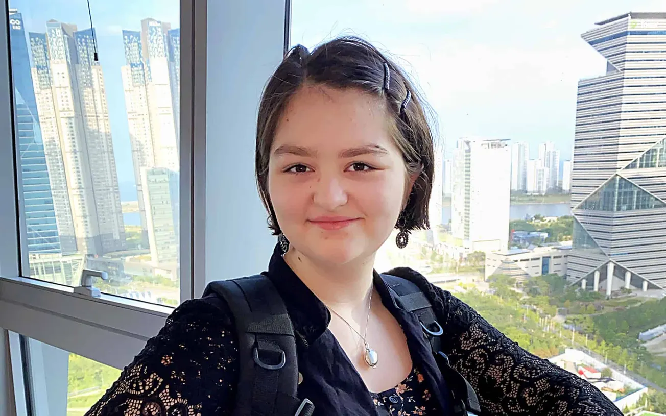Young woman with short dark hair and earrings smiling in front of a cityscape with modern skyscrapers during daytime.