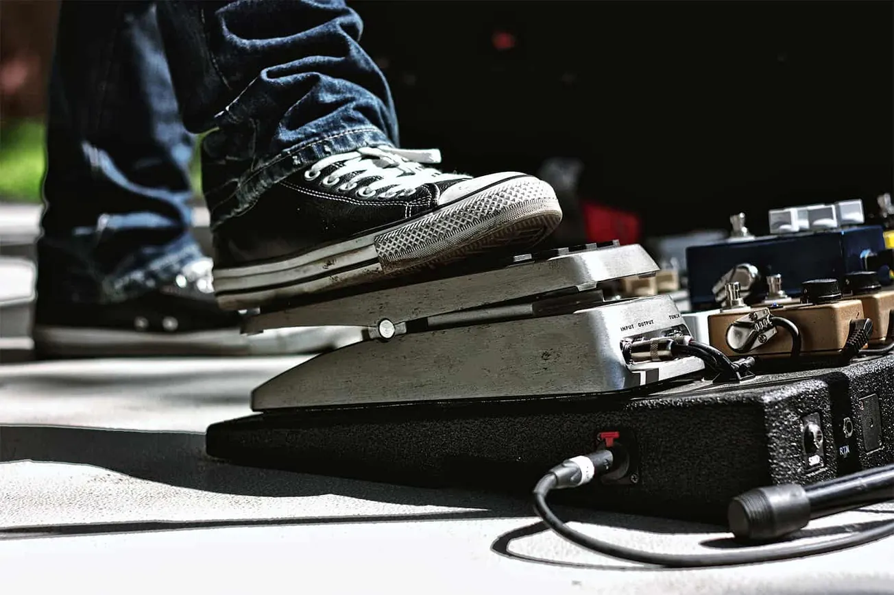 Foot of a musician wearing black Converse sneakers pressing on a guitar effects pedal with multiple pedals in the background,