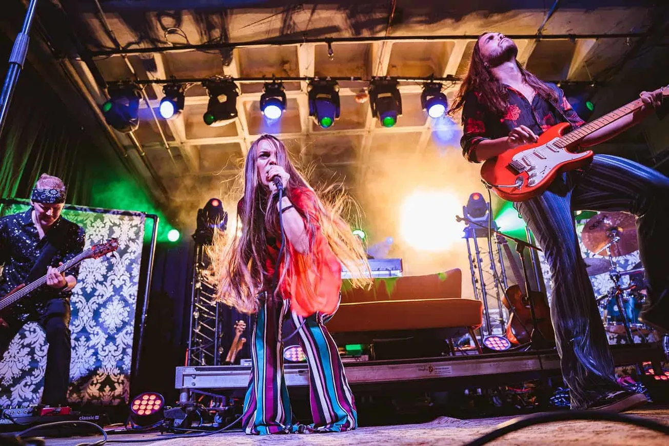 Vibrant live music performance featuring female lead singer and guitarists on stage with colorful lighting and dynamic stage