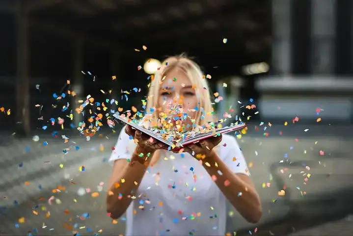 Vibrant woman celebrating with confetti and holding a book outdoors, symbolizing creativity, excitement, and entertainment.