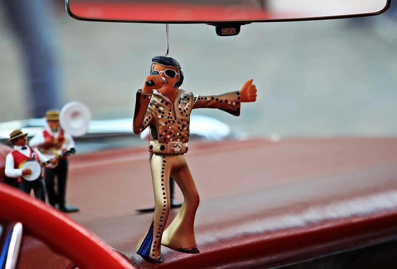 Retro Elvis Presley air freshener hanging from car rearview mirror with mini band figurines in the background.