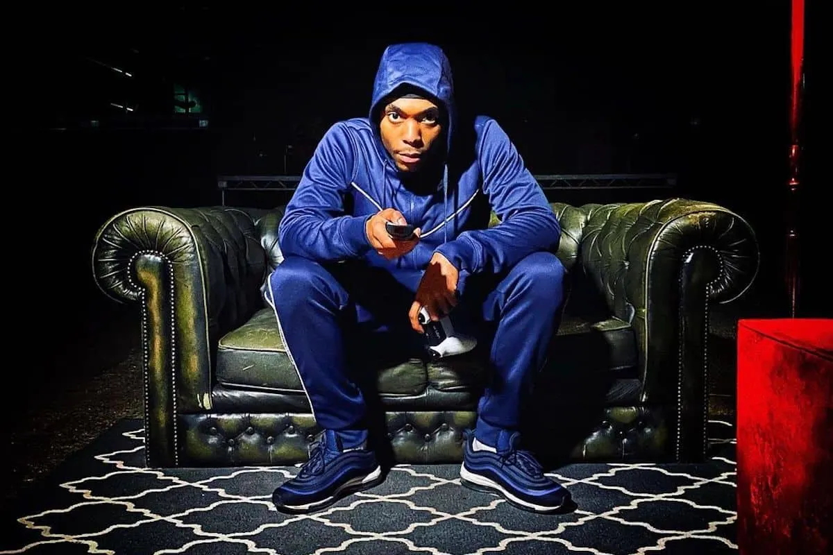 Young man in a blue hoodie and tracksuit sitting on a vintage leather sofa, holding gaming controllers and a phone, in a dark