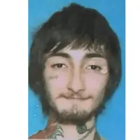 A young man with dark hair, facial hair, and tattoos on his neck, smiling calmly against a blue background, representing mode