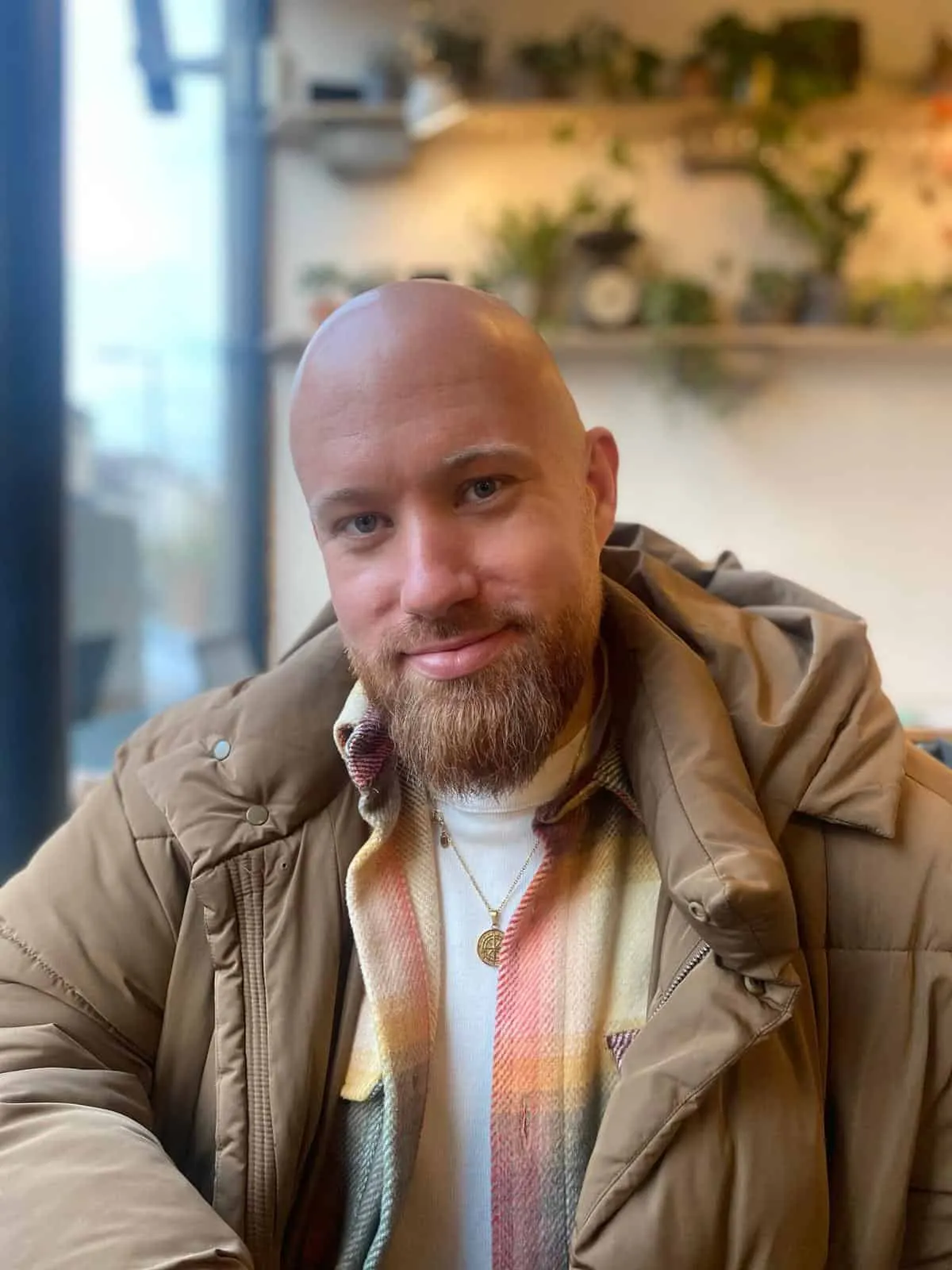 Bald man with a beard smiling in a cozy café, wearing a beige jacket and a plaid scarf, with a background featuring plants an