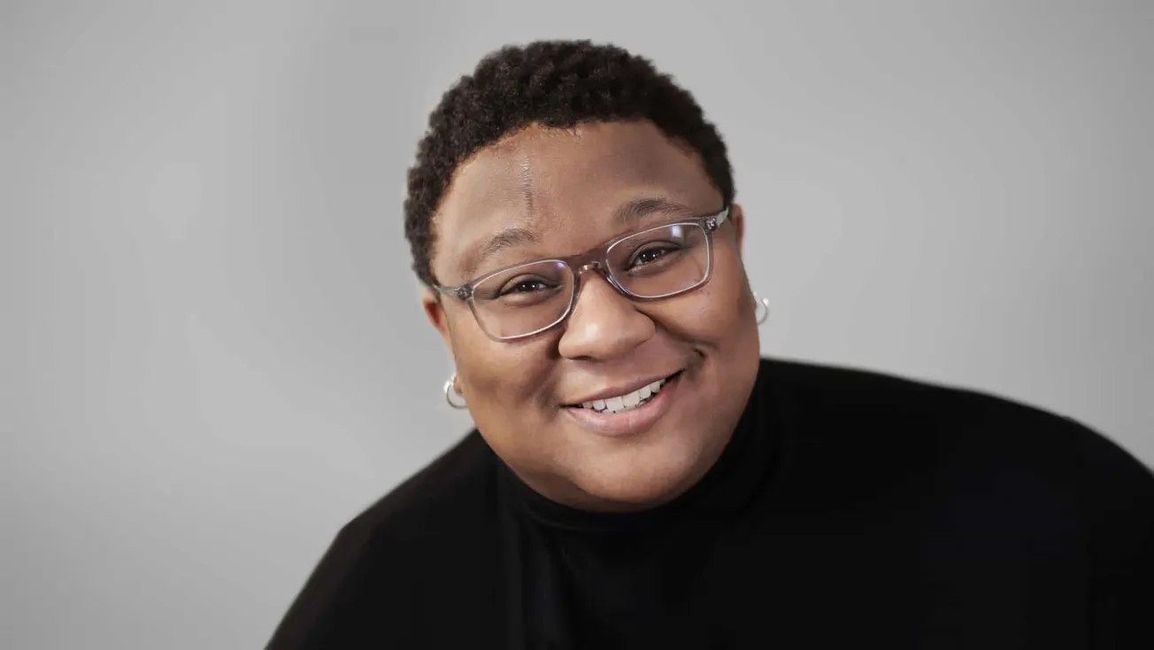 Smiling African American woman wearing glasses and earrings, close-up portrait against gray background.