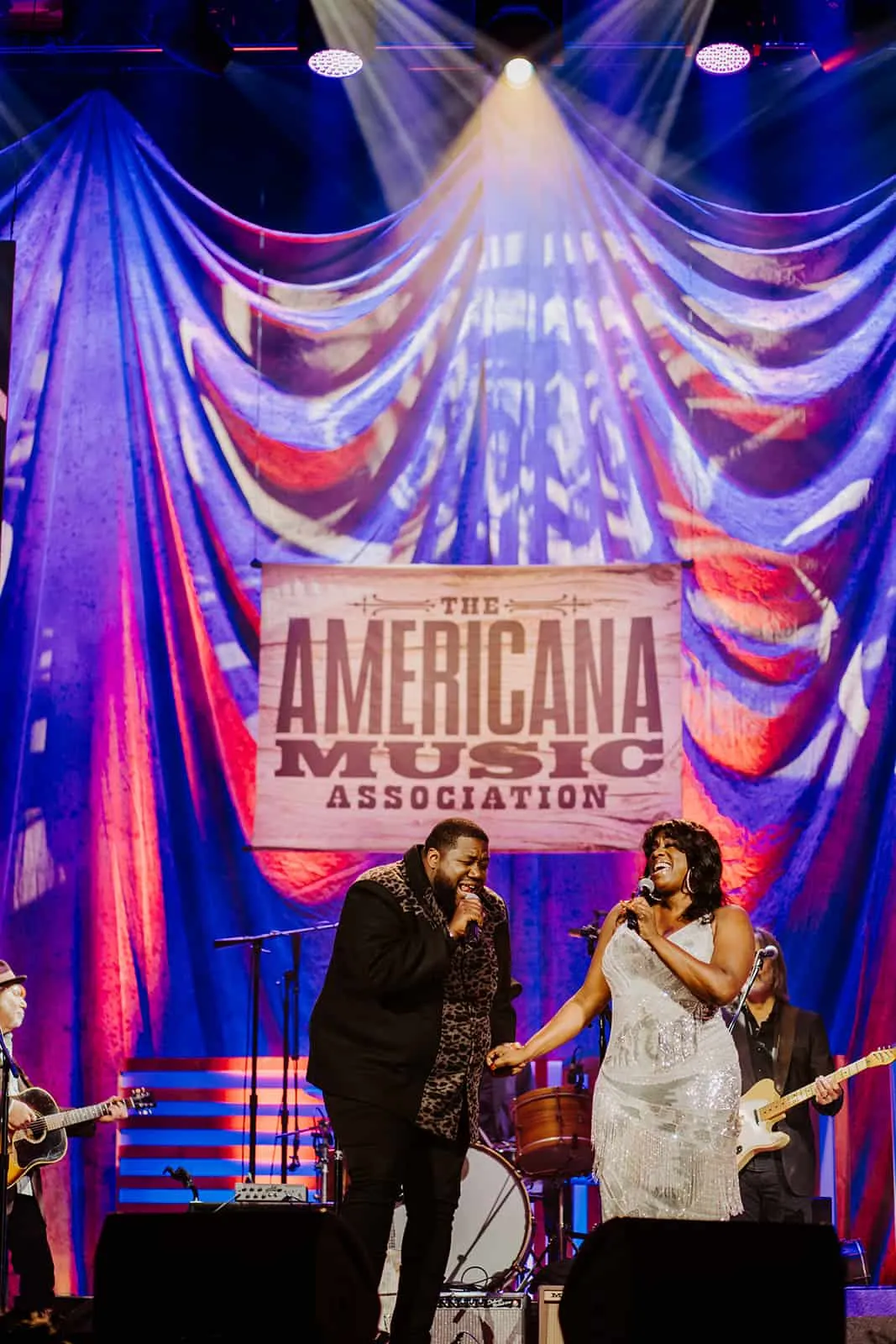 Performers singing live at the Americana Music Association concert with vibrant stage lighting and a dramatic backdrop.