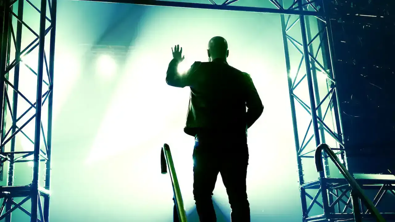 Bald man in black jacket waving on stage with green lighting and metal truss structures at a concert or music event.