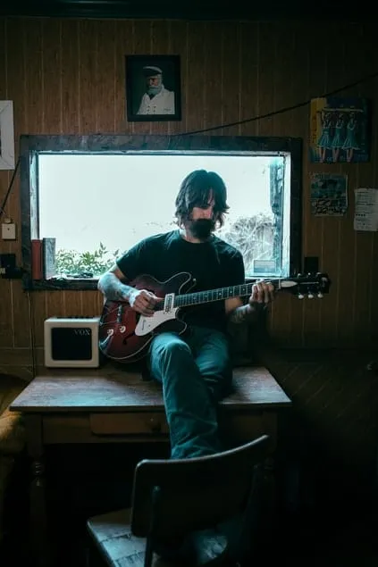 Man playing electric guitar in cozy room with wood-paneled walls and art posters, natural light from window, music studio atm