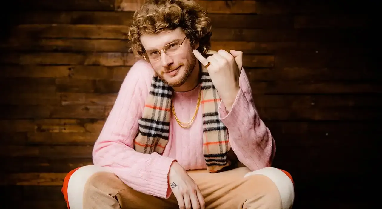 Young man with curly hair and glasses, wearing a pink sweater and a Burberry plaid scarf, sitting in front of a wooden backgr