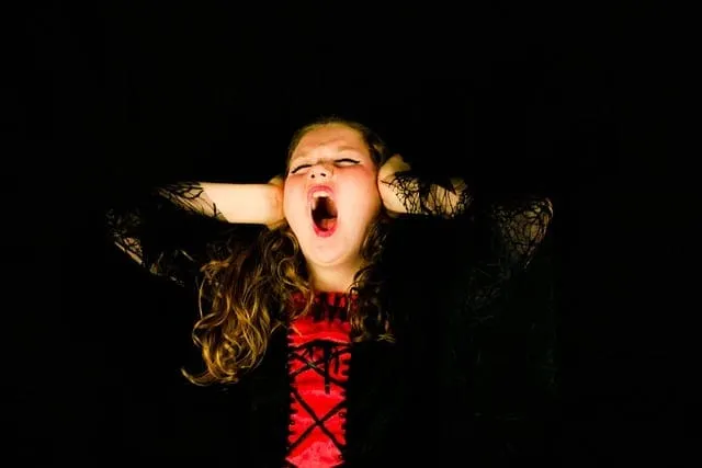 A young girl with curly hair covering her ears, screaming or shouting with a distressed expression against a black background