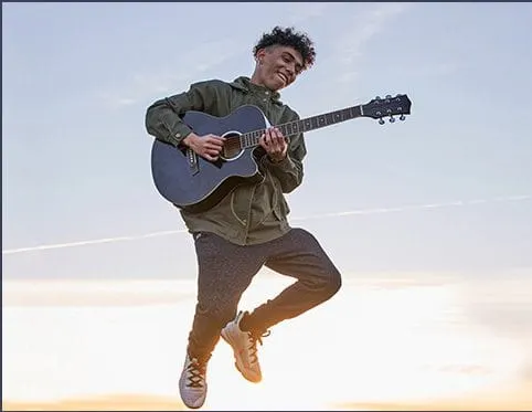 A young man jumping outdoors while playing an acoustic guitar during sunset, showcasing musical creativity and outdoor perfor