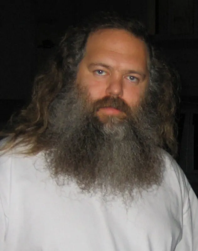 Long-haired man with a full beard and serious expression in a casual white shirt, indoor setting.