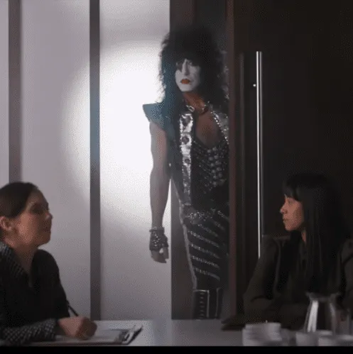 A mime artist dressed as a rock star peeking through a conference room door while two women sit at a table, engaging in a dis