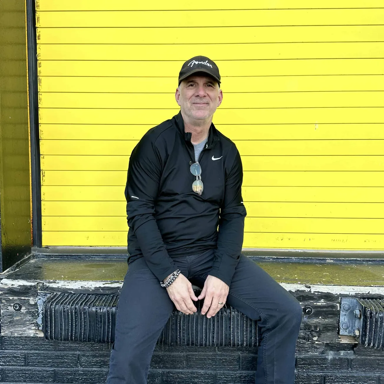Bright yellow wall with black horizontal lines and a man in black Nike sportswear sitting on a weathered wooden bench, casual