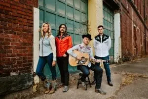 Urban band of four people with one playing guitar, standing outdoors near a brick building, casual street style music scene,