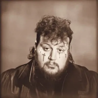 A man with tattoos on his face, curly hair, and a leather jacket, posing against a plain background.