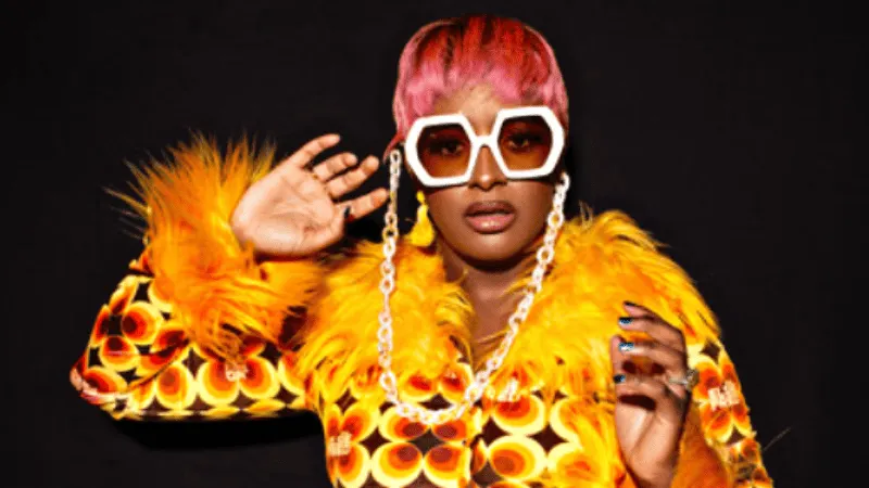 Bold woman with pink hair wearing oversized white sunglasses and a vibrant orange and yellow patterned fur coat, striking a s